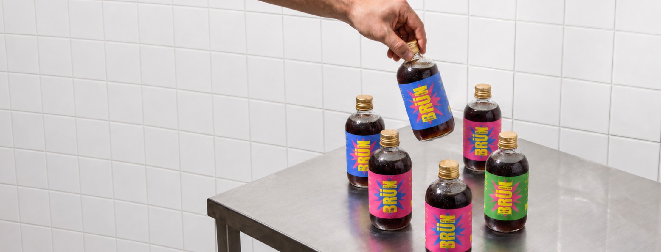 Hand placing a BRÜN cold brew bottle among five others on a steel table